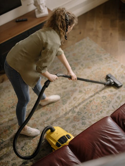 A yellow canister vacuum cleaner with a black hose and metal wand attachment is resting on a beige patterned area rug in a room with wooden flooring. To the left, a black metal and wood media console