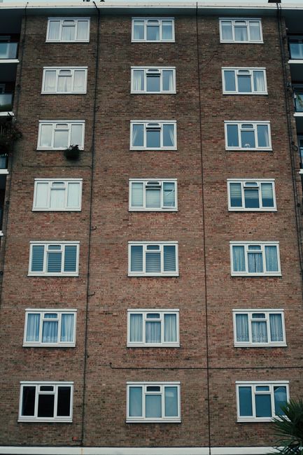 A narrow outdoor balcony running alongside a brick apartment building, featuring a concrete floor and a low brick wall with black metal railings. The balcony has a row of black drain pipes attached to